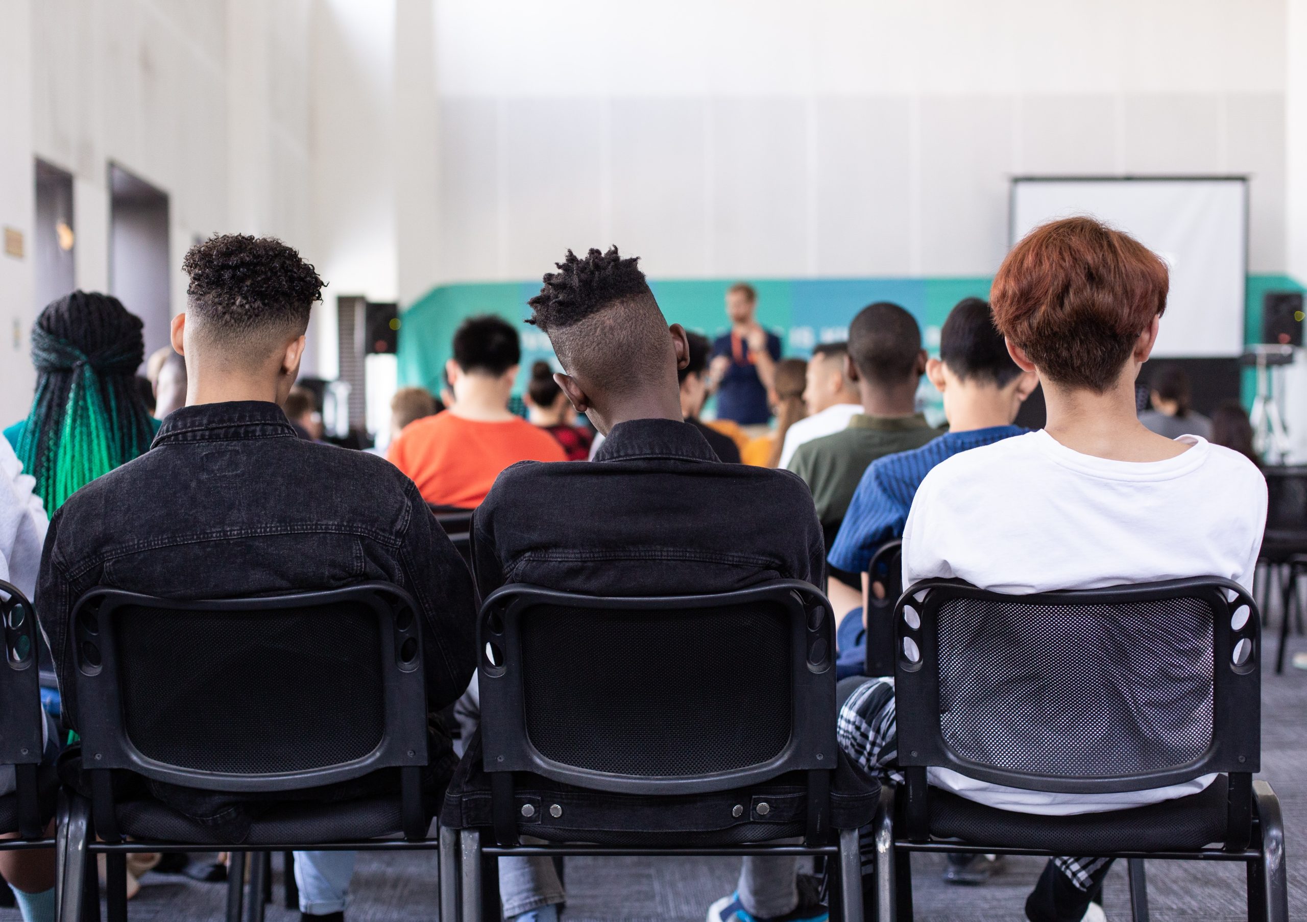 The backs of three people sitting in chairs at the back of a classroom, with postures indicating they are listening to the lecturer
