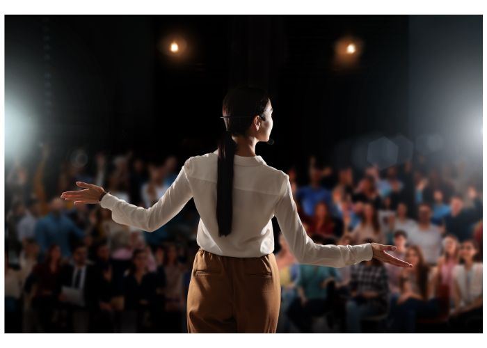 In dimmed lighting and the spotlight on her, a young woman confidently stands in front of an audience with her arms out to her side.