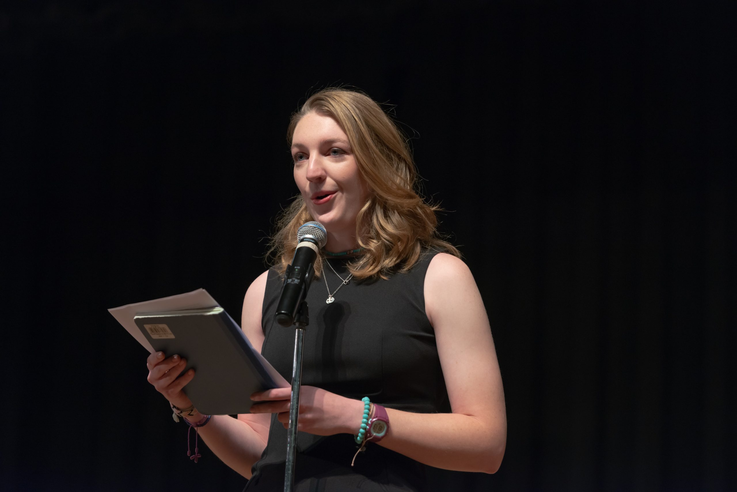 A woman holding a notebook and pieces of paper giving a speech from a microphone.
