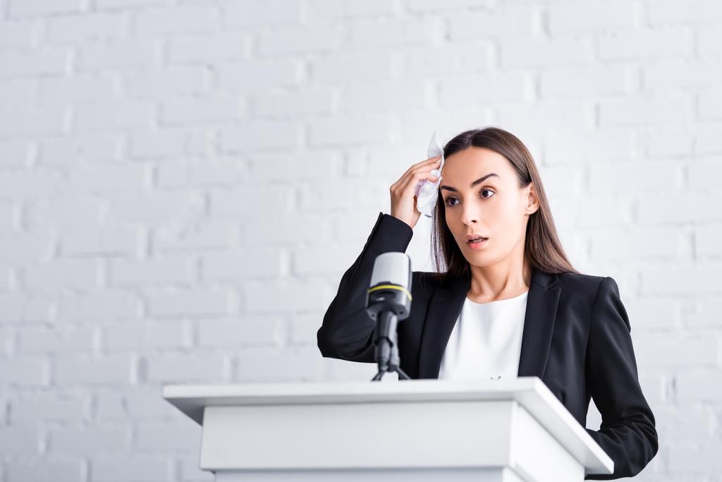 A young female speaker is standing behind a lectern and nervously wiping her brow.