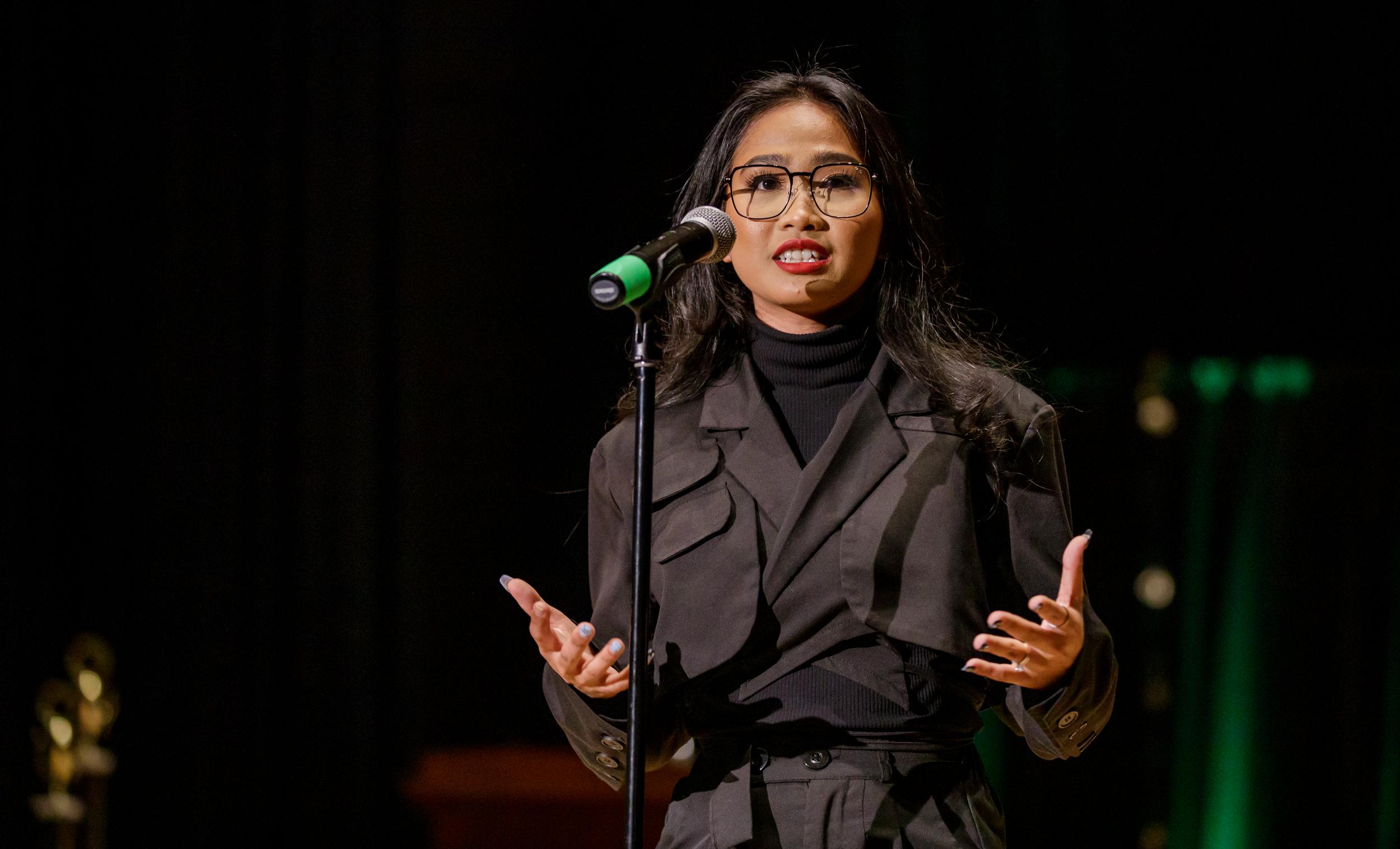 A young woman standing behind a microphone speaks to her audience while simultaneously gesturing passionately.