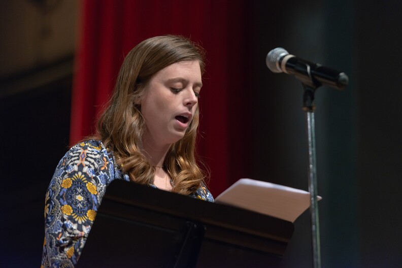 A young woman standing behind a microphone speaks from a manuscript.