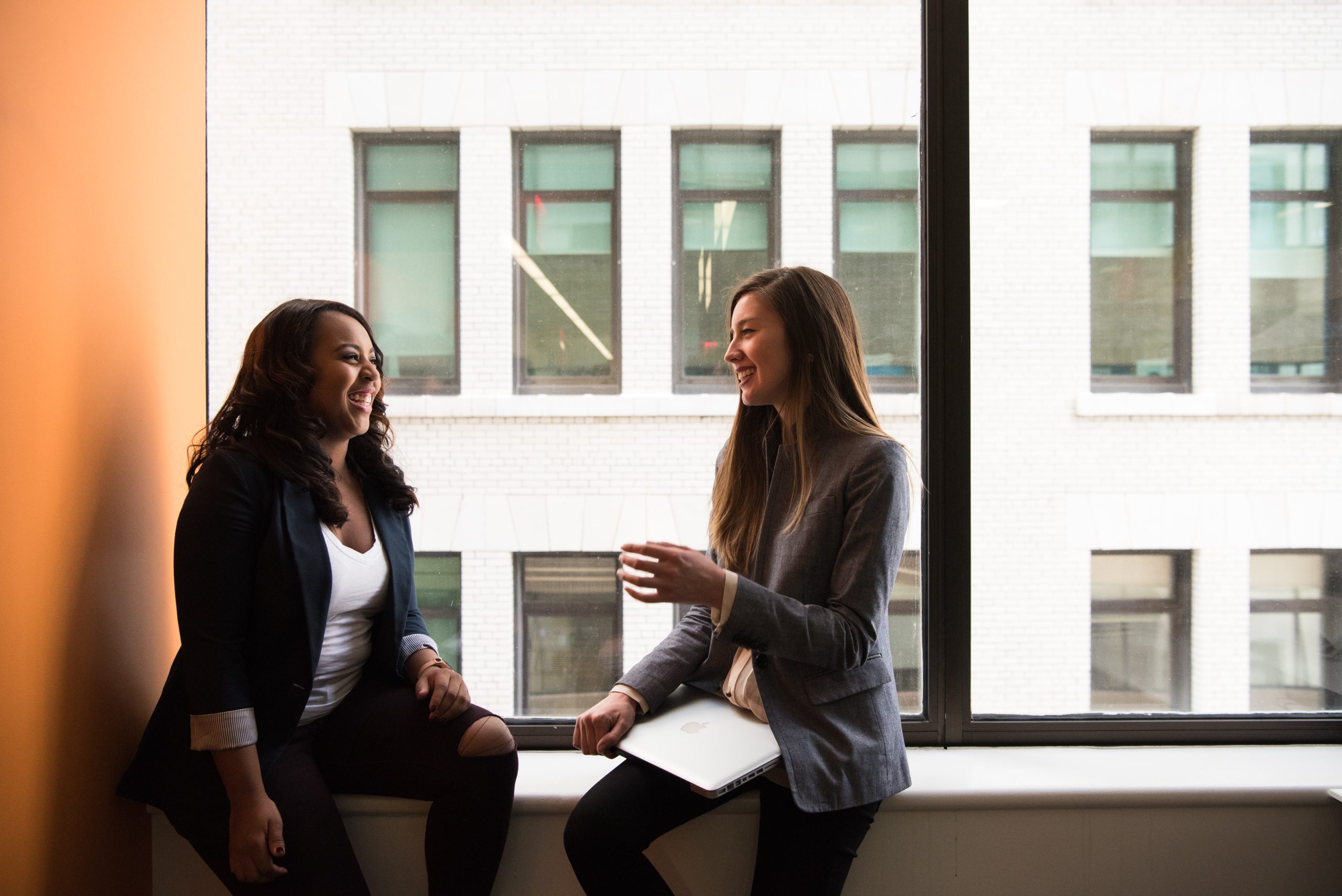 Two women hold a conversation while sitting in a large window sill in a mulitple story building