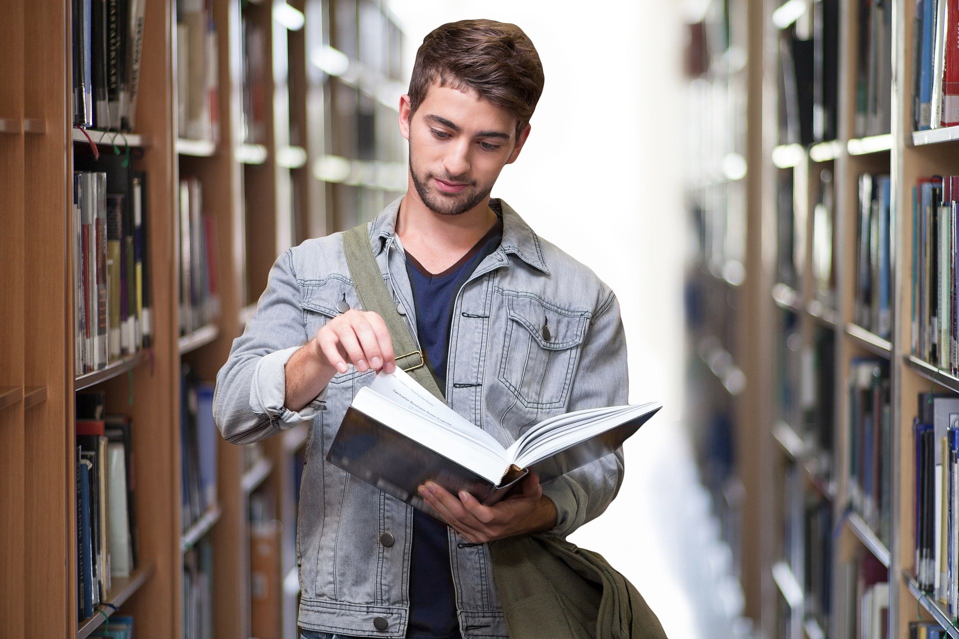 A man standing between bookshelves while holding a book and reading it.