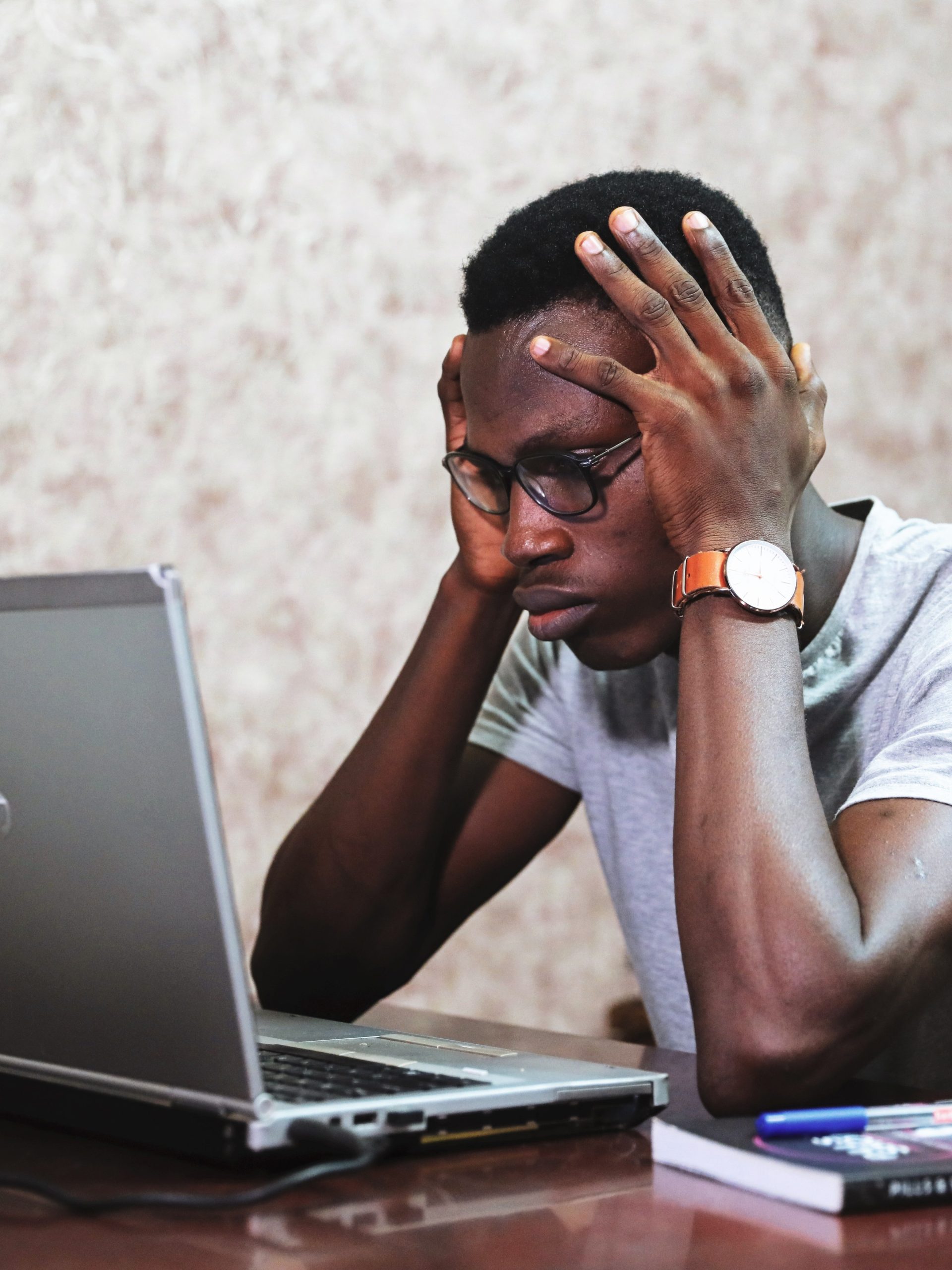 A man with glasses presses both hands to his head while using a laptop
