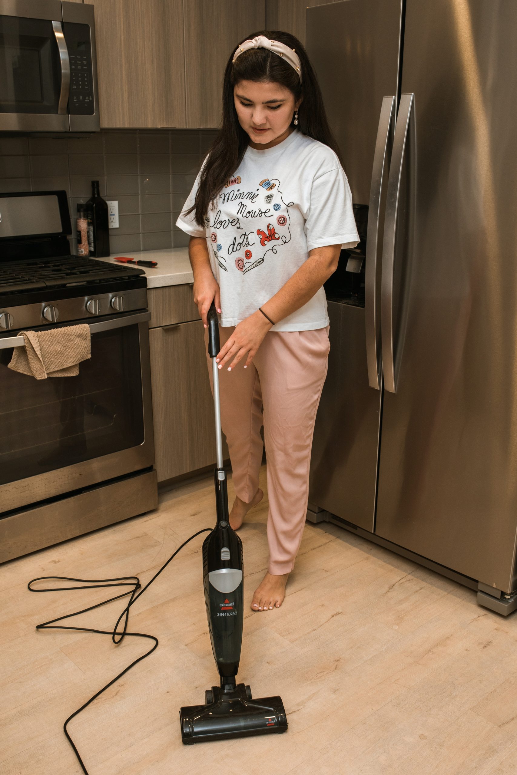 A college aged woman runs a stick vacuum in a kitchen.